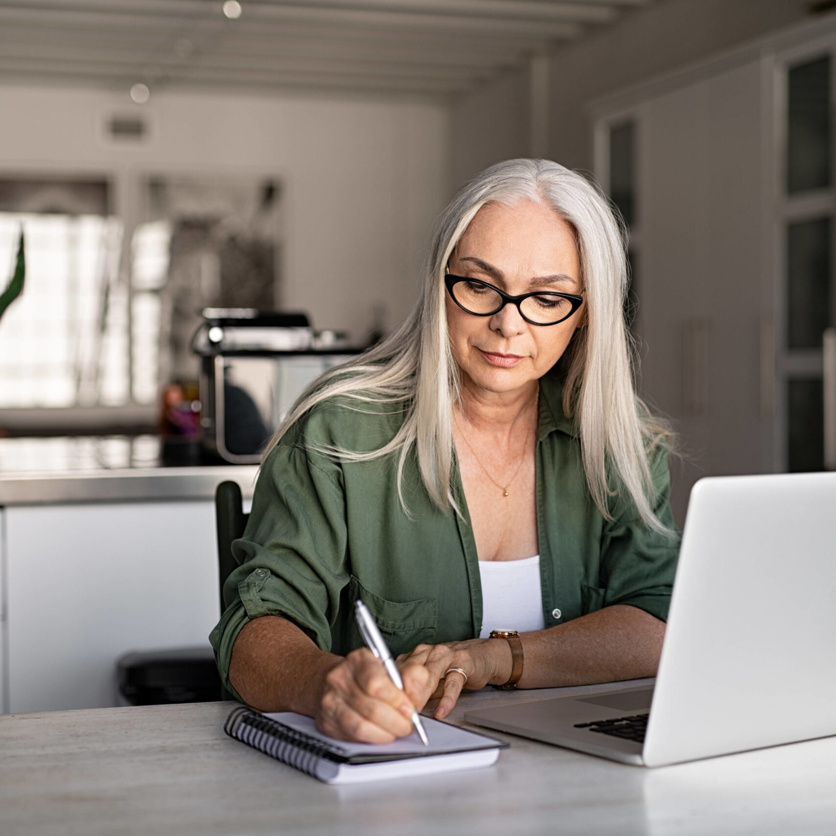 Senior stylish woman taking notes in notebook while using laptop at home. Old freelancer writing details on book while working on laptop in living room. Focused cool lady writing notary in notepad.