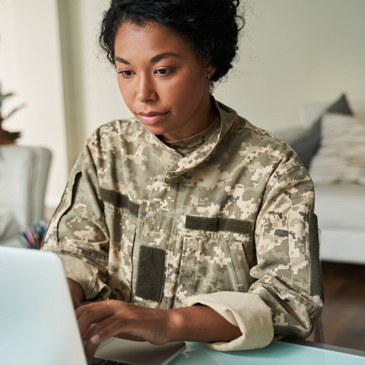 Focused black woman using laptop at table. Concept of modern woman. Female soldier wear camouflage uniform. After war rehabilitation. Blurred background of little boy. Interior of modern apartment