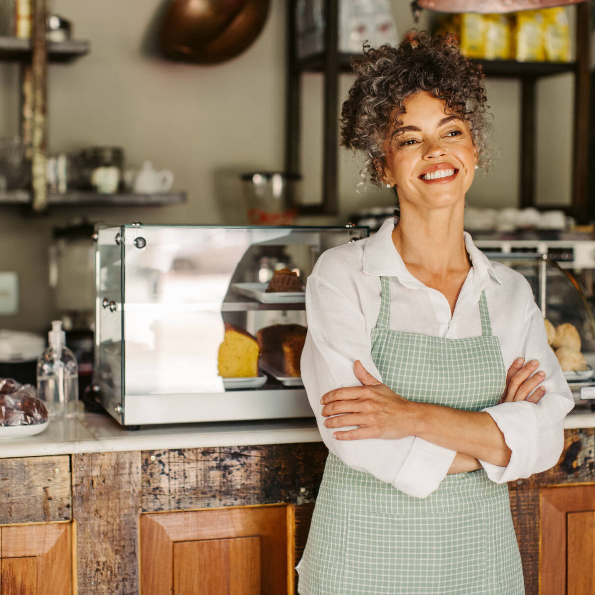 Cheerful business owner smiling while standing with her arms crossed. Successful mature businesswoman feeling confident while wearing an apron in front of her coffee shop during the day.