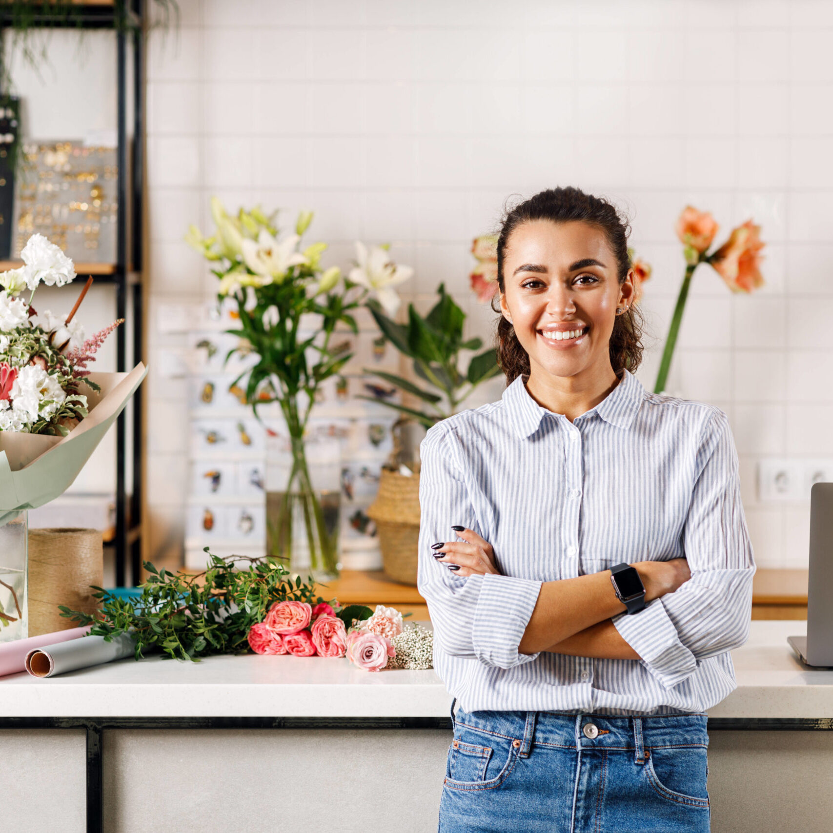 Confident female florist standing with her arms crossed and looking at camera. Happy flower shop owner standing at counter.