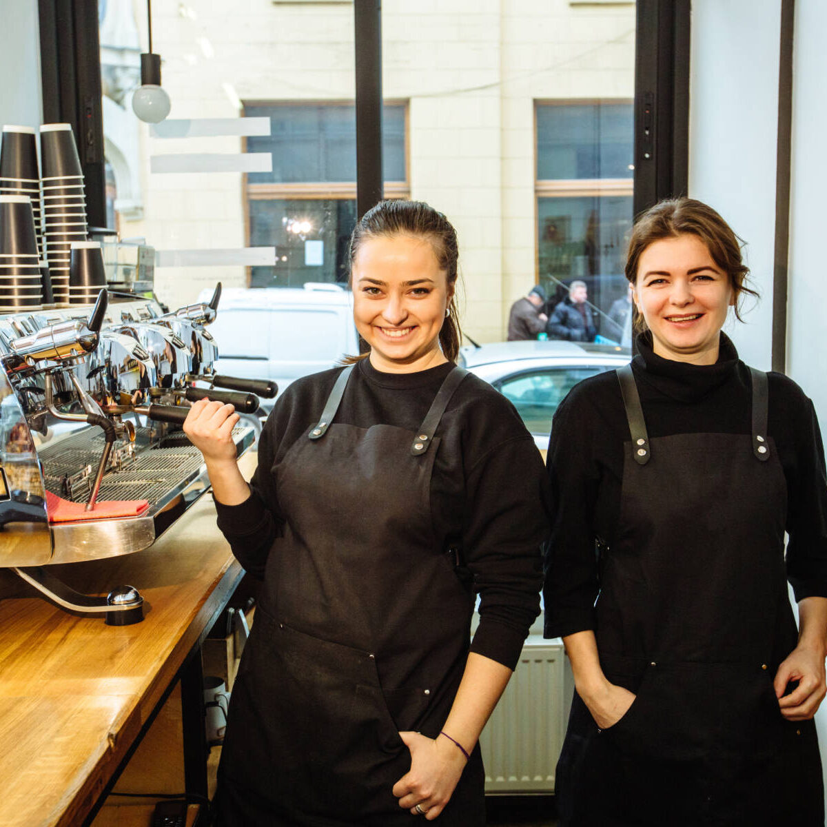 Two laughing real people female baristas in black uniform laughing, standing together in their workplace. Business partners stand behind the counter at coffee shop. Coworking concept