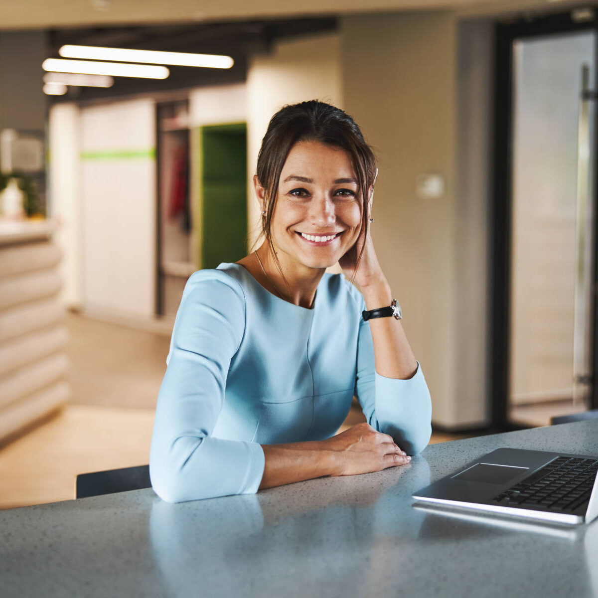 Happy cheerful business lady in blue formal clothes sitting at the work desk while looking at the photo camera in bright modern office