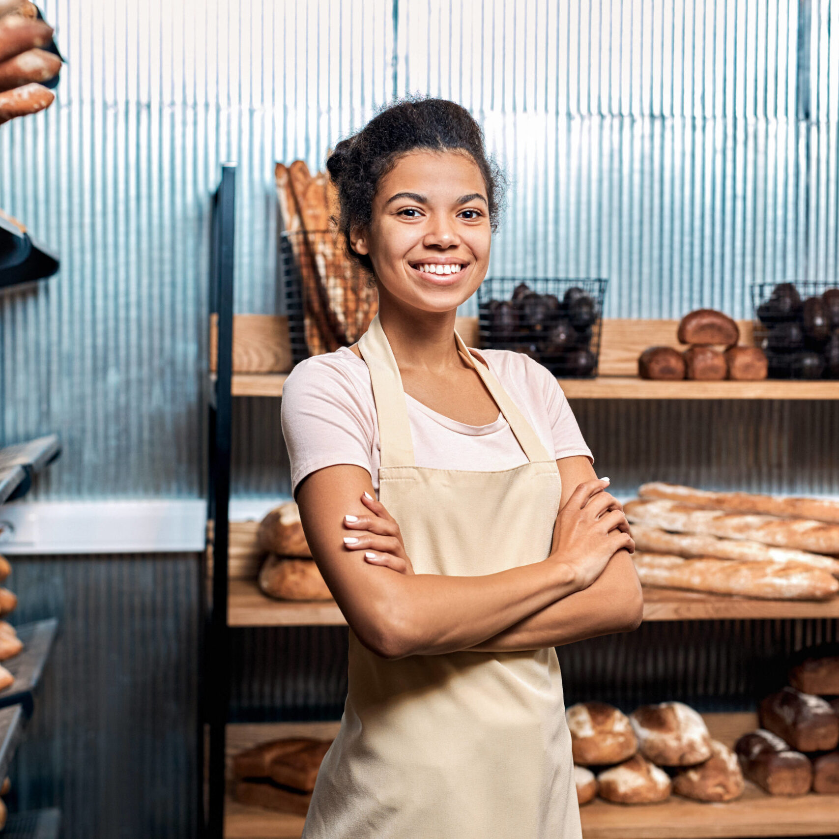 Family business concept. Confident and happy young adult woman working in small bakery, standing near fresh wholegrain bread crossed hands on chest