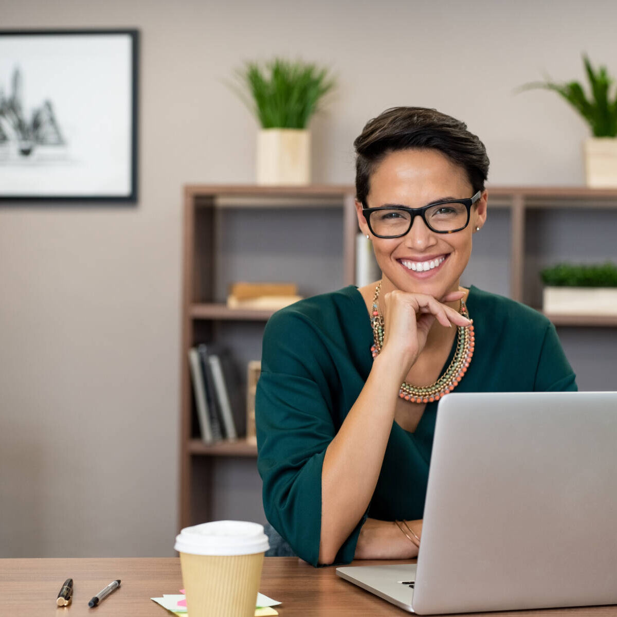 Portrait of happy casual businesswoman sitting at her desk in office. Cheerful latin woman working on laptop and looking at camera. Young fashionable girl wearing eyeglasses sitting at creative agency.